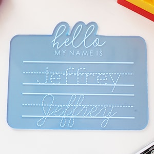 Könnte beinhalten: Ein hellblaues Namensschild aus Kunststoff mit den Worten "hello my name is" und dem Namen "Jeffrey", der in gepunkteten Linien zum Nachzeichnen geschrieben ist.