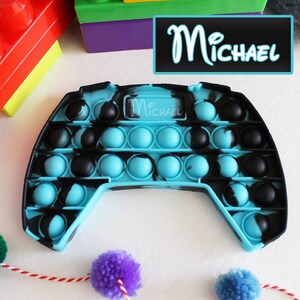 Puede incluir: Un juguete pop it fidget azul y negro con forma de mando de videojuego. El juguete tiene el nombre "Michael" escrito en él.