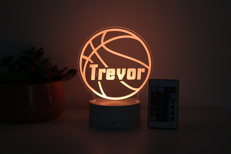 Pu&ograve; includere: Una luce notturna a tema basket con il nome "Trevor" illuminato in bianco. La luce ha un design a palla da basket e poggia su una base bianca. Un telecomando con opzioni di colore &egrave; accanto alla luce.
