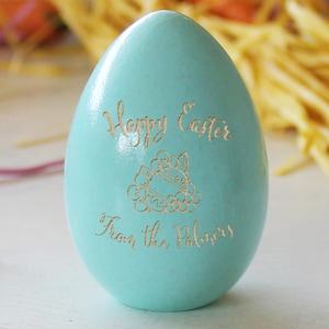 Puede incluir: Un huevo de Pascua azul claro con el texto "Feliz Pascua de los Palmers" y un pequeño diseño grabado de un huevo de Pascua con flores.