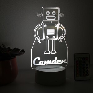 May include: A white robot night light with the name "Camden" underneath. The light is on a white base and comes with a remote control.