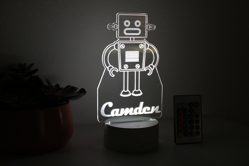 May include: A white robot night light with the name "Camden" underneath. The light is on a white base and comes with a remote control.