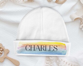 Pode incluir: Gorrinho branco para bebé com uma faixa de arco-íris de aquarela e o nome "CHARLES" em texto preto.