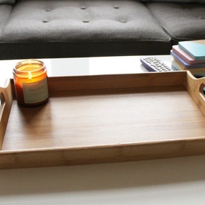 May include: A wooden serving tray with two handles on the sides. The tray is empty and is sitting on a white surface. There is a lit candle and a remote control on the surface next to the tray.