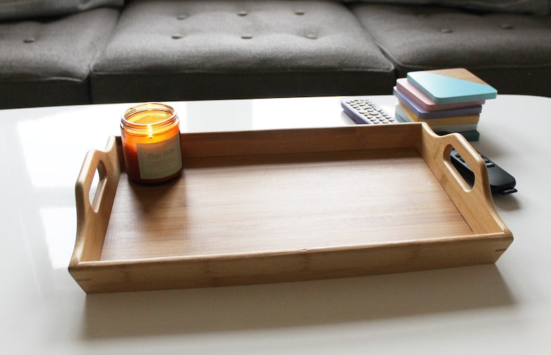 May include: A wooden serving tray with two handles on the sides. The tray is empty and is sitting on a white surface. There is a lit candle and a remote control on the surface next to the tray.