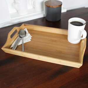 May include: A rectangular bamboo serving tray with two handles. The tray is empty except for three silver spoons and a white napkin. A white mug with a black handle is sitting on the tray.