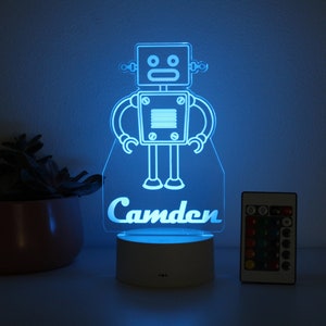 May include: A blue robot night light with the name "Camden" printed below. The light is on a white base and has a remote control.