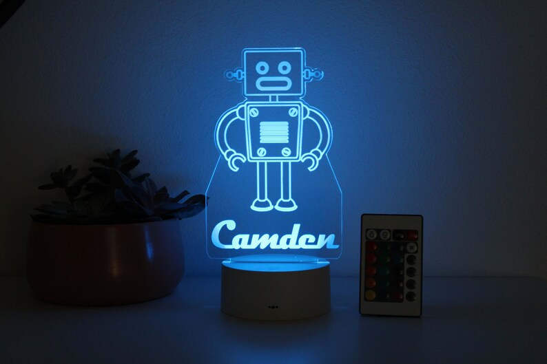 May include: A blue robot night light with the name "Camden" printed below. The light is on a white base and has a remote control.