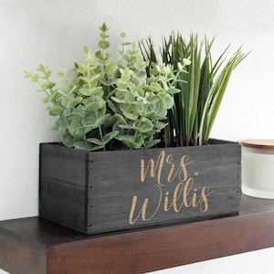 May include: A rectangular, dark gray wooden planter box with the name "Mrs. Willis" in a cursive font. The box is filled with green artificial plants, including eucalyptus and tall grass. The planter sits on a dark brown shelf.