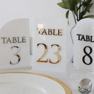 May include: Three white acrylic table number signs with gold, silver, and black numbers. The signs read "Table 23", "Table 3", and "Table 8".