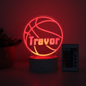 Pu&ograve; includere: Luce notturna a LED a forma di pallone da basket rosso con il nome "Trevor" illuminato. La luce &egrave; su una base bianca, con un telecomando accanto. Adatta per una cameretta.