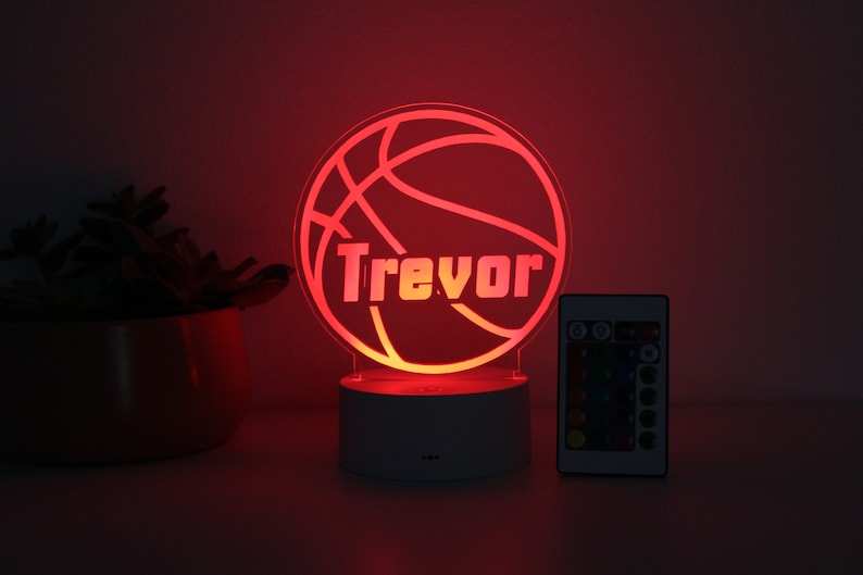 Pu&ograve; includere: Luce notturna a LED a forma di pallone da basket rosso con il nome "Trevor" illuminato. La luce &egrave; su una base bianca, con un telecomando accanto. Adatta per una cameretta.