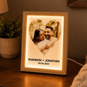 May include: A rectangular light-up frame with a light wood finish featuring a heart-shaped photograph of two people embracing. The text SHANNON + JONATHAN and the date 05.06.2023 are printed in black below the heart. The frame emits a warm glow.