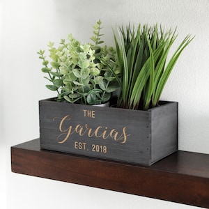 May include: A dark gray wooden planter box with the name "The Garcias" and "EST. 2018" in gold lettering. The box is filled with green plants and sits on a dark brown shelf against a white wall. A decorative home accent.