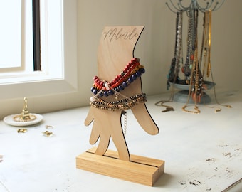 May include: Wooden hand-shaped jewelry stand with the name "Melinda" engraved on the arm. The stand is holding a variety of colorful beaded bracelets.