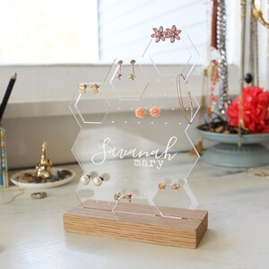 Op de afbeelding: Een transparante, zeshoekige oorbellenhouder van acryl met een houten basis. De houder heeft meerdere gaten voor oorbellen en een ruimte voor een armband. De houder is gepersonaliseerd met de naam "Savannah mary".