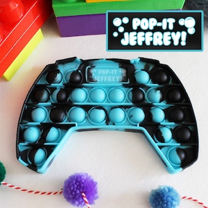 Puede incluir: Un juguete pop-it anti estrés azul y negro con forma de mando de videojuego. El juguete tiene las palabras "Pop-it Jeffrey!" escritas en él.