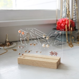 May include: A clear acrylic earring holder with a wooden base. The holder has a geometric design and is filled with various earrings, including pearl, flower, and stud earrings.