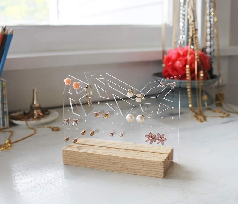 May include: A clear acrylic earring holder with a wooden base. The holder has a geometric design and is filled with various earrings, including pearl, flower, and stud earrings.