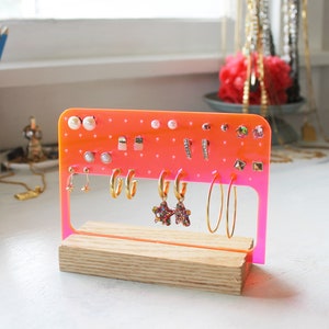 May include: A pink acrylic earring holder with a wooden base. The holder has multiple holes for earrings and is filled with various earrings, including studs, hoops, and drop earrings.