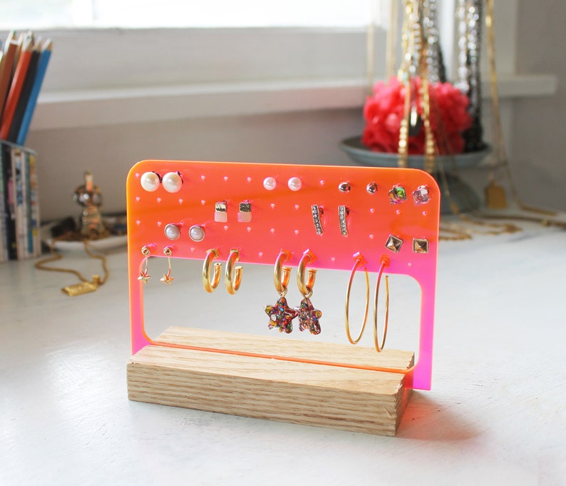 May include: A pink acrylic earring holder with a wooden base. The holder has multiple holes for earrings and is filled with various earrings, including studs, hoops, and drop earrings.