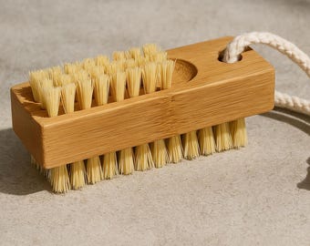 May include: A natural bamboo nail brush with light-colored bristles. The rectangular brush has a curved indentation and a loop of off-white rope for hanging. The bristles are densely packed on both sides of the brush.