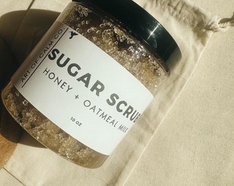 May include: A glass jar filled with a honey and oatmeal milk sugar scrub. The label reads "Art of Calm Co. Sugar Scrub Honey + Oatmeal Milk 10 oz."