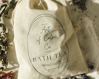 May include: A beige canvas bag labeled "Art of Calm Co BATH TEA" with a green oval design and a drawing of a bathtub. The bag is on a white marble plate, surrounded by dried flowers, herbs, and bath salts, creating a spa-like aesthetic.