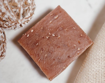 May include: A close-up of a rectangular bar of brown soap with a grainy texture. The soap is sitting on a white surface with a piece of beige burlap fabric in the background.