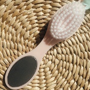 Puede incluir: Un cepillo para pies rosa con un cepillo de cerdas blancas en un extremo y un exfoliante plateado en el otro. El cepillo está sobre una estera de paja tejida.
