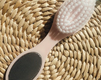 May include: A pink foot brush with a white bristle brush on one end and a silver scrub on the other end. The brush is lying on a woven straw mat.