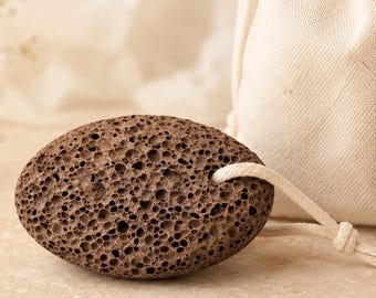 May include: An oval-shaped dark brown pumice stone with a porous, textured surface and a light-colored cord loop attached, accompanied by a small, off-white drawstring pouch made of woven fabric with matching cotton drawstrings.