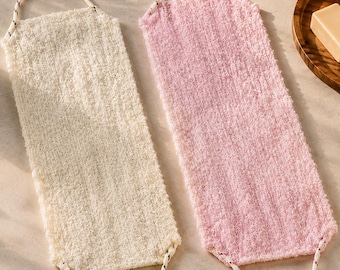 May include: Two textured bath scrubbers, one cream and one pink, with rope handles. The scrubbers are rectangular and have a loop for hanging. Other bath accessories, including a wooden soap dish, are in the background.