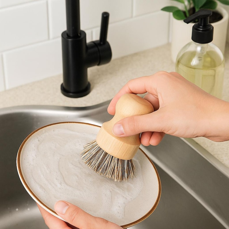 Puede incluir: Un cepillo de madera para lavar platos con cerdas naturales frota un plato blanco con borde marr&oacute;n. El plato est&aacute; en un fregadero de acero inoxidable. Un grifo negro y un dispensador de jab&oacute;n est&aacute;n en el fondo.