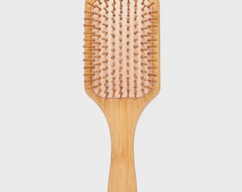 May include: A rectangular wooden hairbrush with a handle. The brush has a light brown wooden frame with a light pink cushion and brown bristles. The handle is rounded at the end. The brush is designed for hair care.