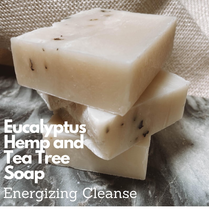 May include: Three bars of white soap stacked on top of each other. The soap has a speckled appearance. The text 'Eucalyptus Hemp and Tea Tree Soap Energizing Cleanse' is printed below the soap bars.