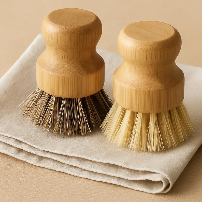 Puede incluir: Dos cepillos de cocina de madera con cerdas naturales. Uno tiene cerdas marrones, el otro cerdas de color claro. Los cepillos tienen un mango redondeado y est&aacute;n sobre un pa&ntilde;o plegado de color claro.