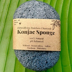 Puede incluir: Una esponja konjac de carbón de bambú negra con una etiqueta de papel marrón que dice "Detoxifying Bamboo Charcoal Konjac Sponge 100% Natural pH Balanced Without Preservatives Sulfate Parabens Phthalates".