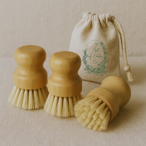 Puede incluir: Tres cepillos de madera para fregar platos con cerdas de color claro y una bolsa con cord&oacute;n a juego. Los cepillos tienen un mango redondeado y son de madera de color claro. La bolsa tiene el texto "Art of Calm Co."