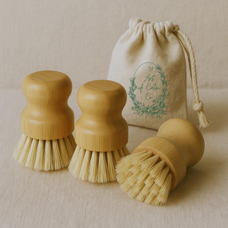 Puede incluir: Tres cepillos de madera para fregar platos con cerdas de color claro y una bolsa con cord&oacute;n a juego. Los cepillos tienen un mango redondeado y son de madera de color claro. La bolsa tiene el texto "Art of Calm Co."