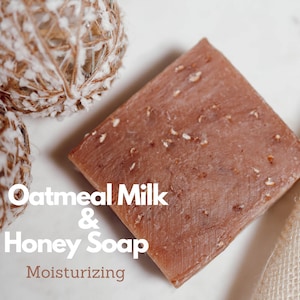 May include: A rectangular bar of brown oatmeal milk and honey soap. The soap has a light brown color and is speckled with small white flecks. The text 'Oatmeal Milk & Honey Soap' is written in white letters below the soap. The text 'Moisturizing' is written in white letters below the text 'Oatmeal Milk & Honey Soap'.