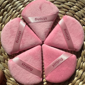 May include: Five pink triangular makeup sponges with a pink ribbon around each one that says "strong" in white lettering.
