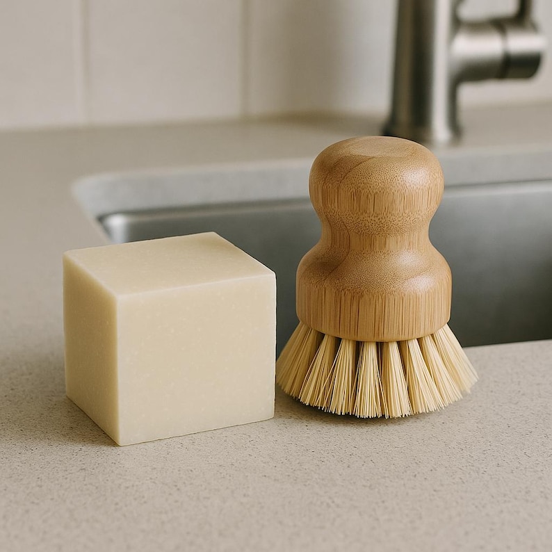 Puede incluir: Un cubo de jab&oacute;n beige claro y un cepillo de cocina de madera con cerdas claras. El cepillo tiene un mango redondeado de bamb&uacute;. Los art&iacute;culos est&aacute;n sobre una encimera clara cerca de un fregadero de acero inoxidable.