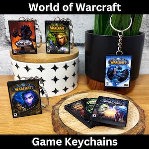 Könnte beinhalten: Schlüsselanhänger mit World of Warcraft-Spielcovern. Die Schlüsselanhänger zeigen verschiedene Spielbox-Art, einschließlich Charaktere und Logos. Die Schlüsselanhänger sind rechteckig mit silbernen Schlüsselringen. Das Bild enthält den Text "World of Warcraft" und "Game Keychains".