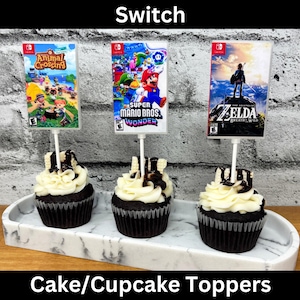 May include: Three chocolate cupcakes with white frosting and chocolate drizzle, each topped with a game cartridge image on a stick. The images are from the Nintendo Switch games Animal Crossing, Super Mario Bros. Wonder, and Zelda.