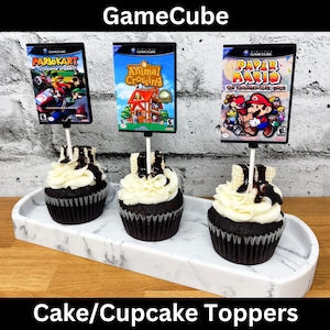 May include: Three cupcakes with white frosting and chocolate drizzle, each topped with a miniature GameCube game case. The games are "Mario Kart: Double Dash", "Animal Crossing", and "Paper Mario". The words "GameCube" and "Cake/Cupcake Toppers" are displayed.