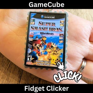 Puede incluir: Un Fidget Clicker con una miniatura de la caja del juego de GameCube para "Super Smash Bros. Melee". La caja del juego tiene un borde gris e incluye la carátula del juego con personajes como Mario y Pikachu. También se ve la palabra "CLICK!".