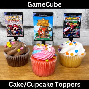 May include: Three cupcakes with GameCube game cover toppers. The toppers display "Paper Mario", "Animal Crossing", and "Mario Kart: Double Dash!!". The cupcakes have colourful frosting and sprinkles. Text at the top says "GameCube" and at the bottom "Cake/Cupcake Toppers".