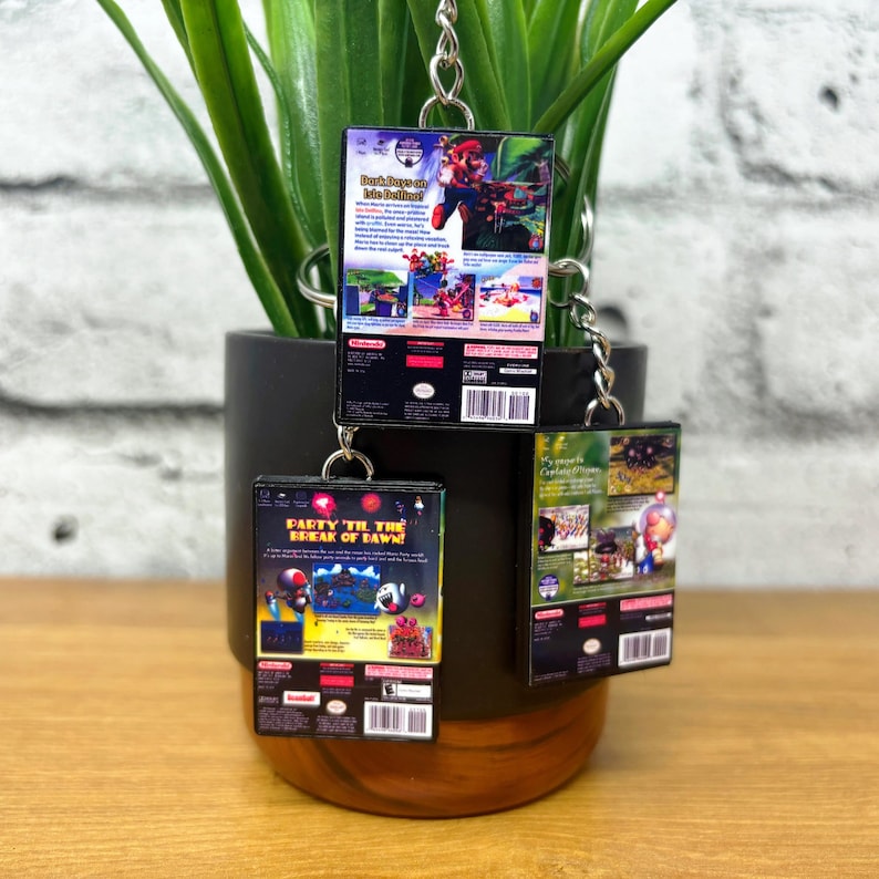 May include: Three miniature video game cases on keychains. The cases are black with colourful images and text. The text on the cases reads "Dark  Dawn", "Party Til The Break of Dawn!" and "My Secret  Campground".