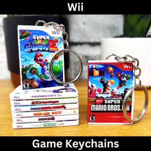 Puede incluir: Llaveros con miniaturas de juegos de Wii. Uno muestra "Super Mario Galaxy 2" y otro "New Super Mario Bros. Wii". También se ve una pila de otras cajas de juegos de Wii. La palabra "Wii" está en la parte superior de la imagen.